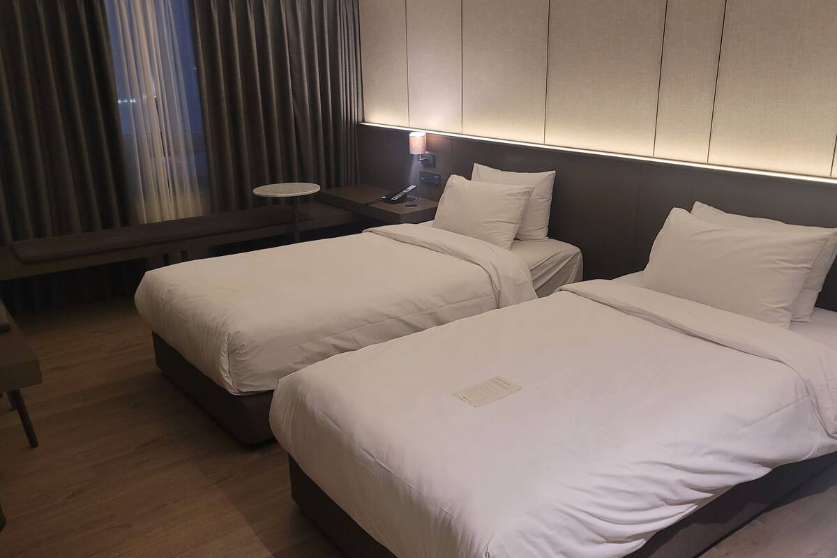 A hotel room with two beds, a desk, and a nightstand with a lamp.