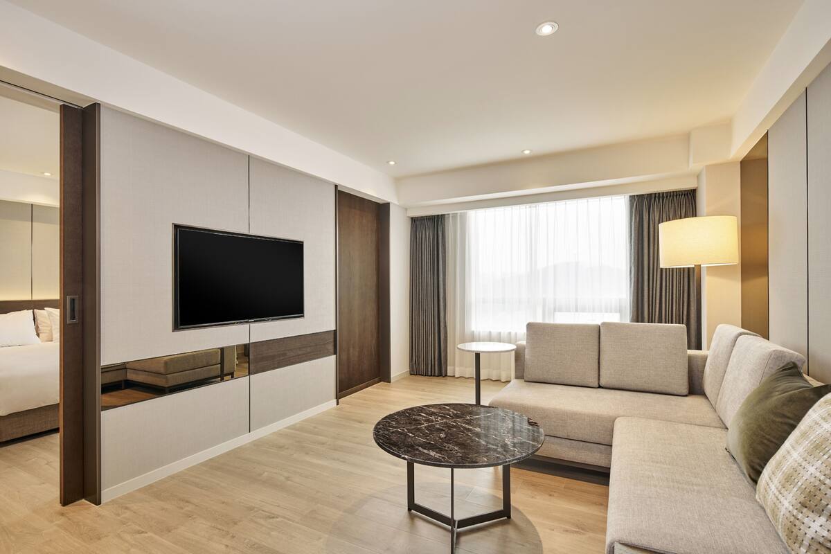 A modern hotel room with a flat-screen TV mounted on the wall, a round coffee table, a sofa, and a bed.