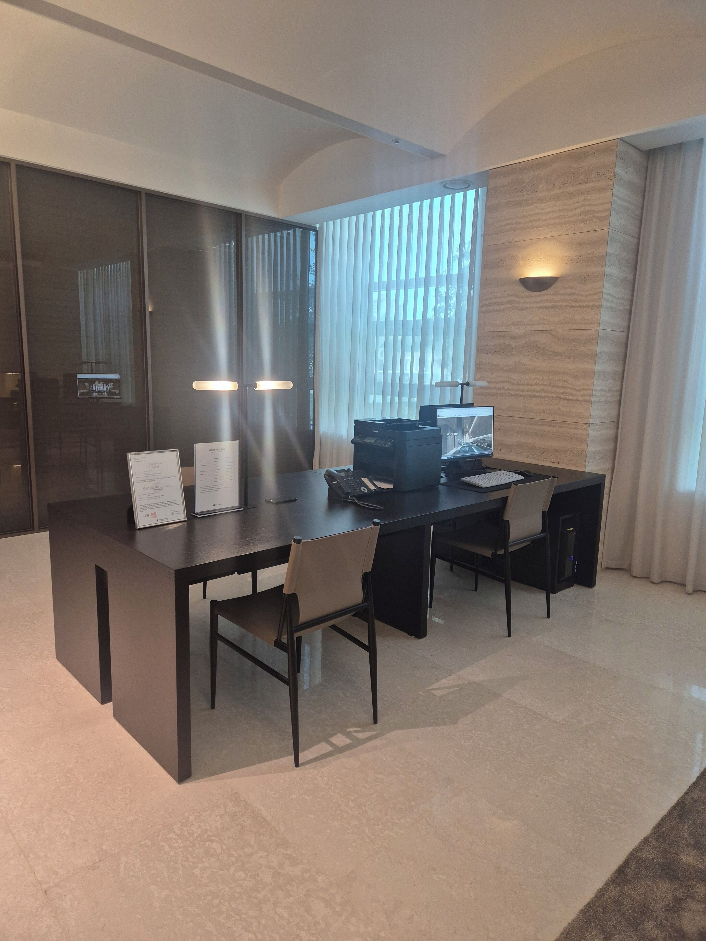 A modern hotel room with a dark wooden desk, two chairs, a laptop, a telephone, and a flat-screen TV.