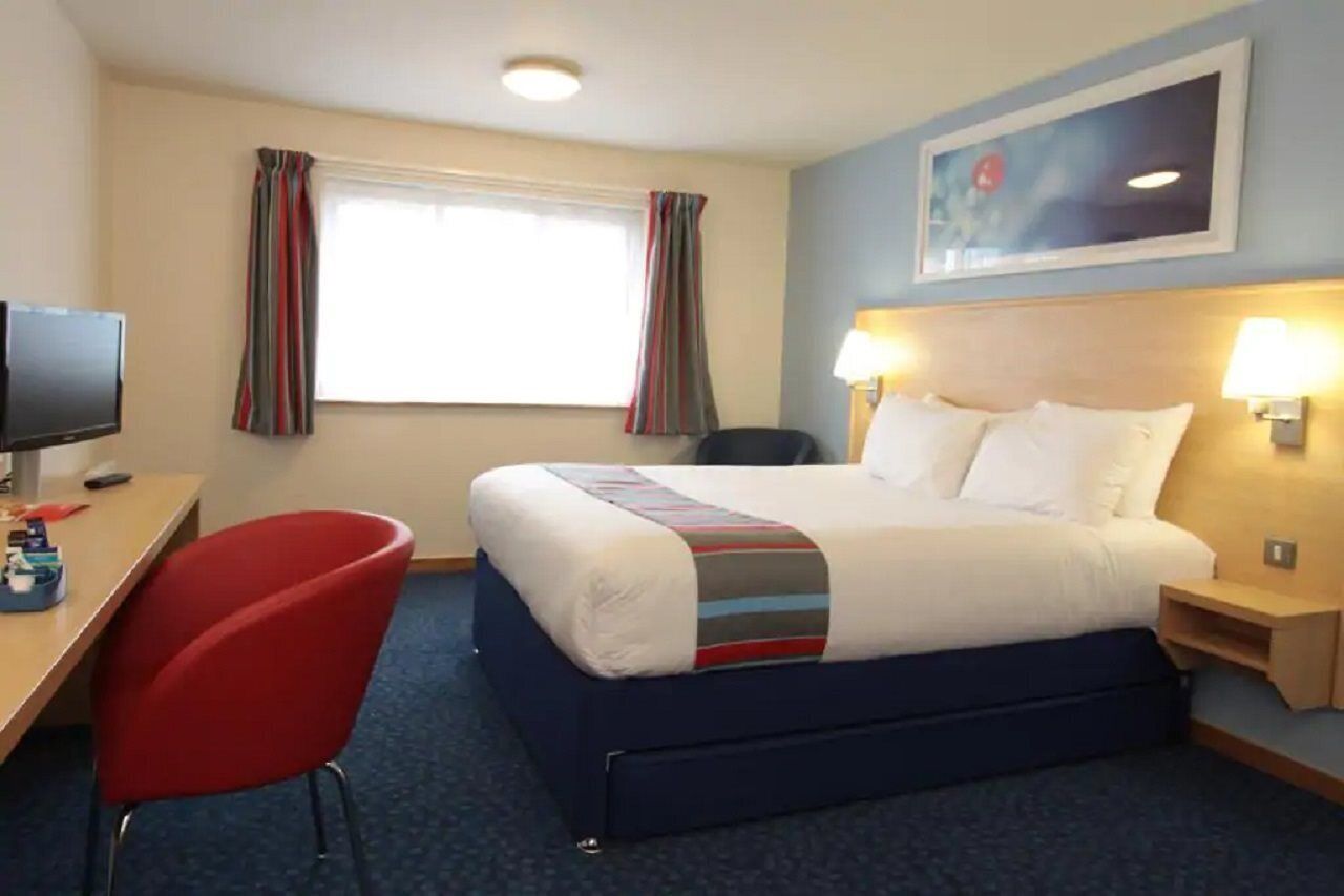 A hotel room with a bed, a desk with a computer, a red chair, and a window with curtains.