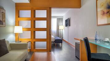 A hotel room with a sofa, a desk with a chair, a TV, a bed, and a glass table with a bottle and cup.