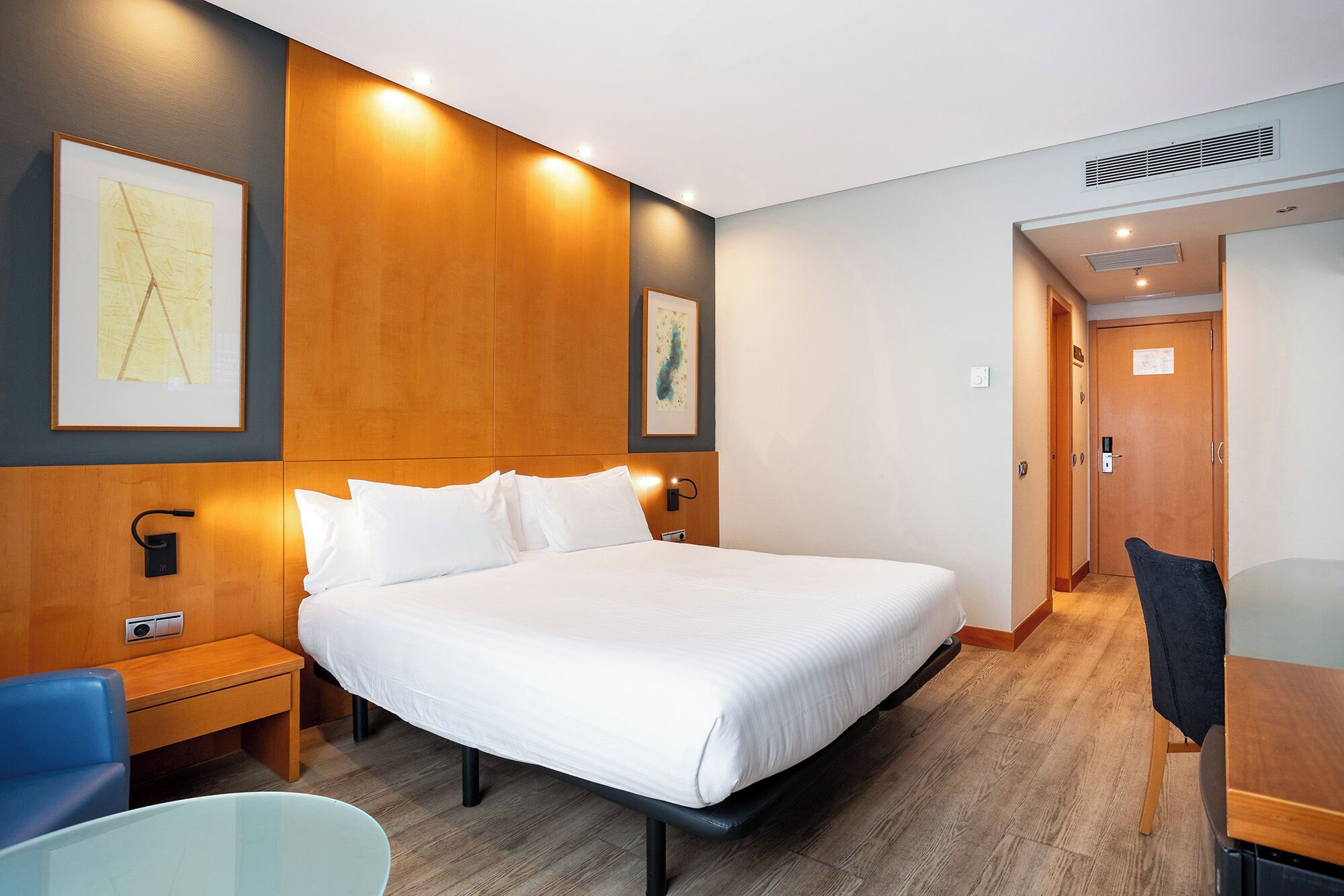 A hotel room with a large bed, a bedside table, a chair, and artwork on the walls.
