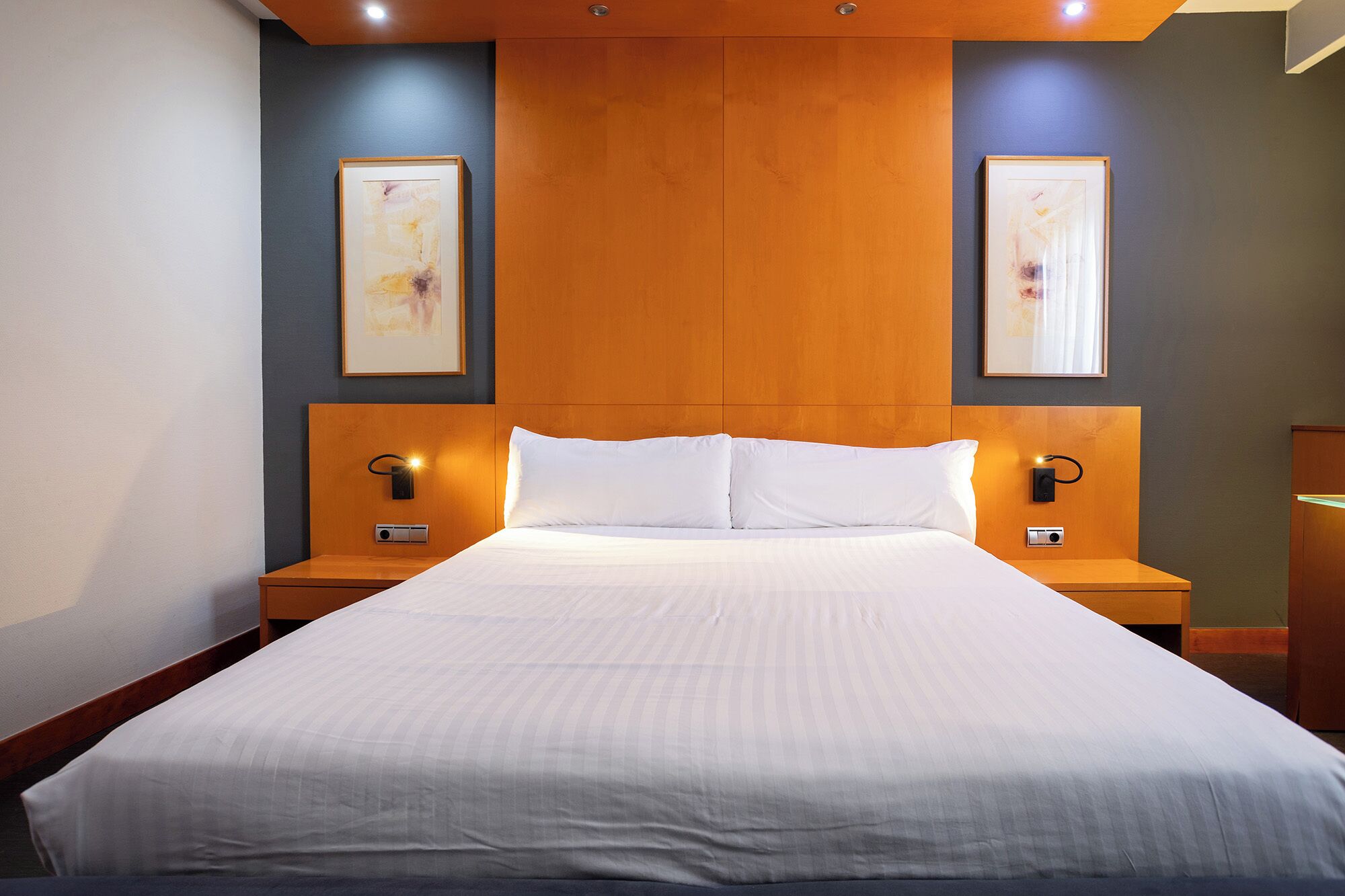 Habitación de hotel con una cama grande, cabecera de madera, dos mesitas de noche con lámparas y dos cuadros en las paredes.