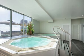Un área de piscina interior moderna con dos jacuzzis, una planta en maceta y vistas de la ciudad a través de grandes ventanales.