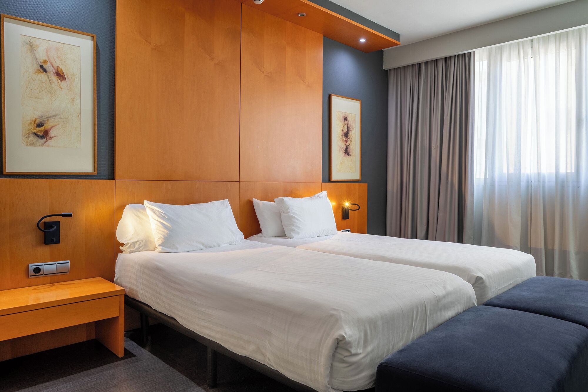 A hotel room with two beds, a bedside table, a bench, and a large window with curtains.