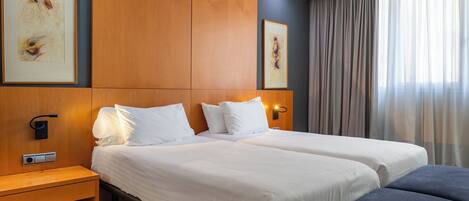 A hotel room with two beds, a bedside table, a bench, and a large window with curtains.