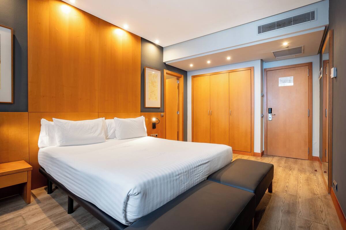 Een moderne hotelkamer met een groot bed, houten lambrisering aan de muren, een bankje aan het voeteneinde van het bed en een deur met een bordje.