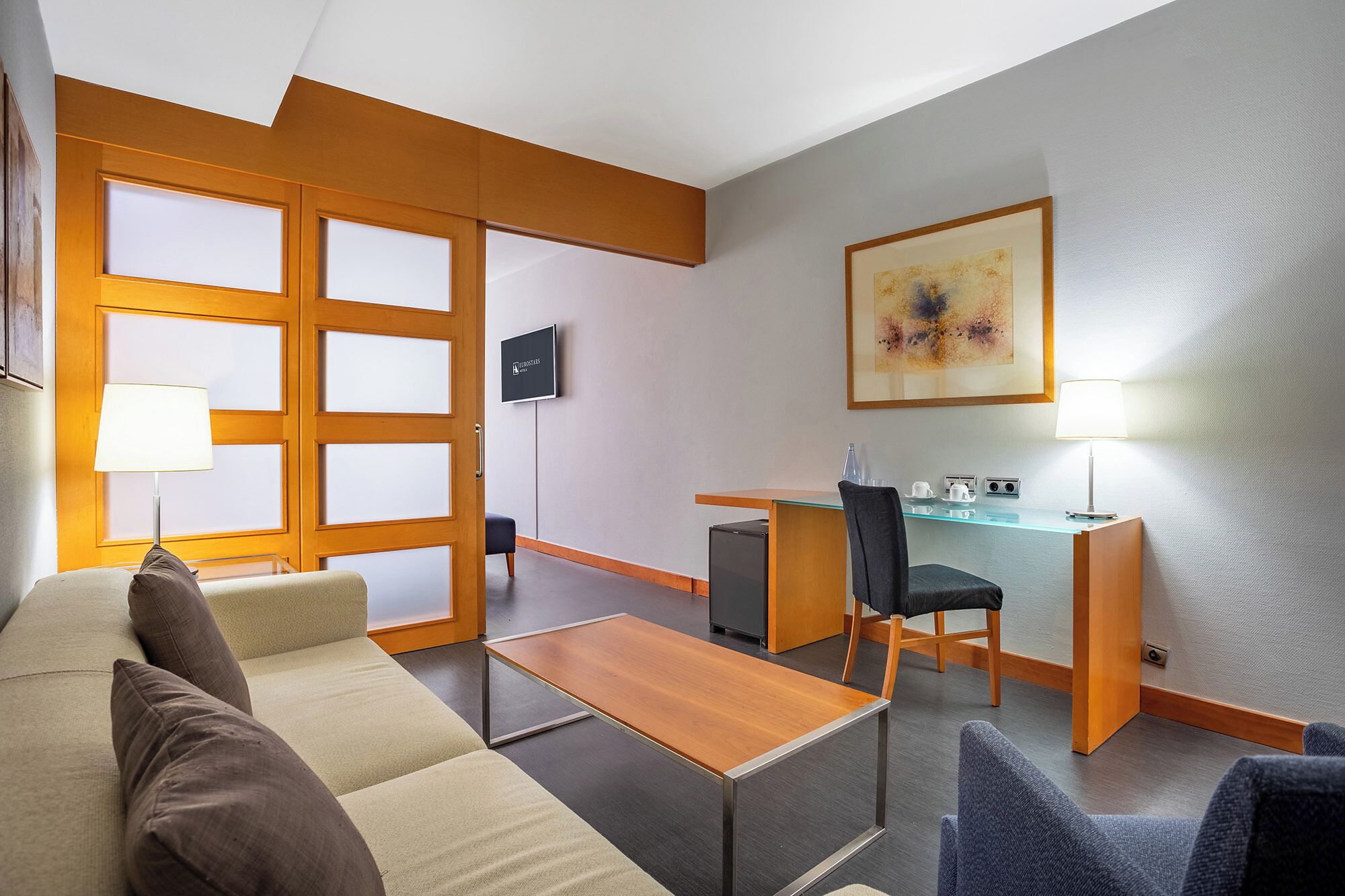 A hotel room with a sofa, a small table, a desk, and a chair. There is a television on the wall and a painting above the desk.