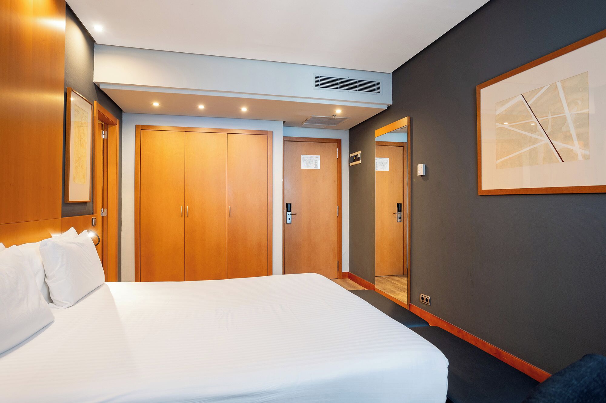 A hotel room with a large bed, wooden wardrobes, a mirror, and a framed abstract artwork on the wall.