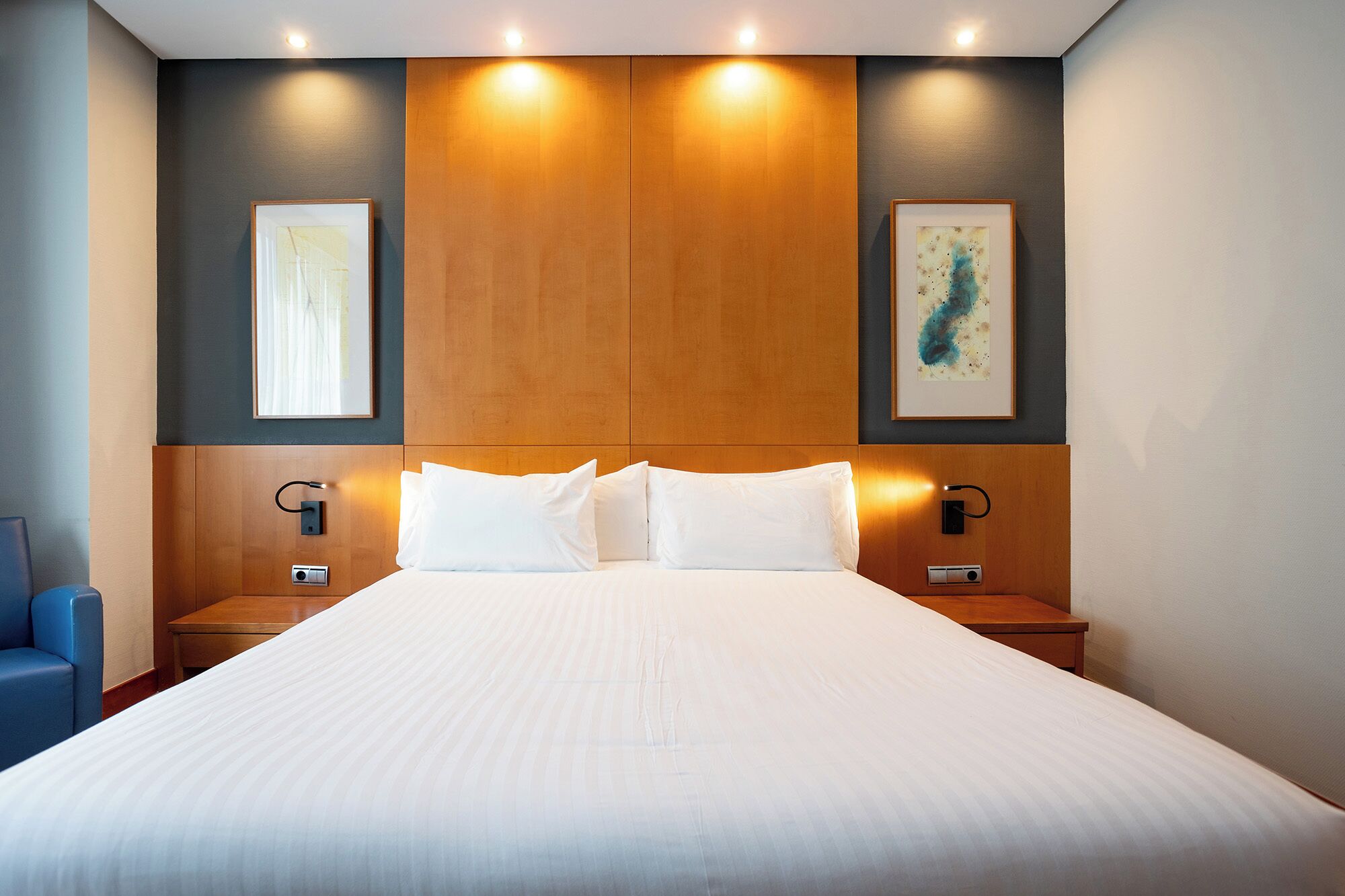 A hotel room with a large bed, two bedside tables, a headboard with built-in lighting, and two framed artworks on the walls.