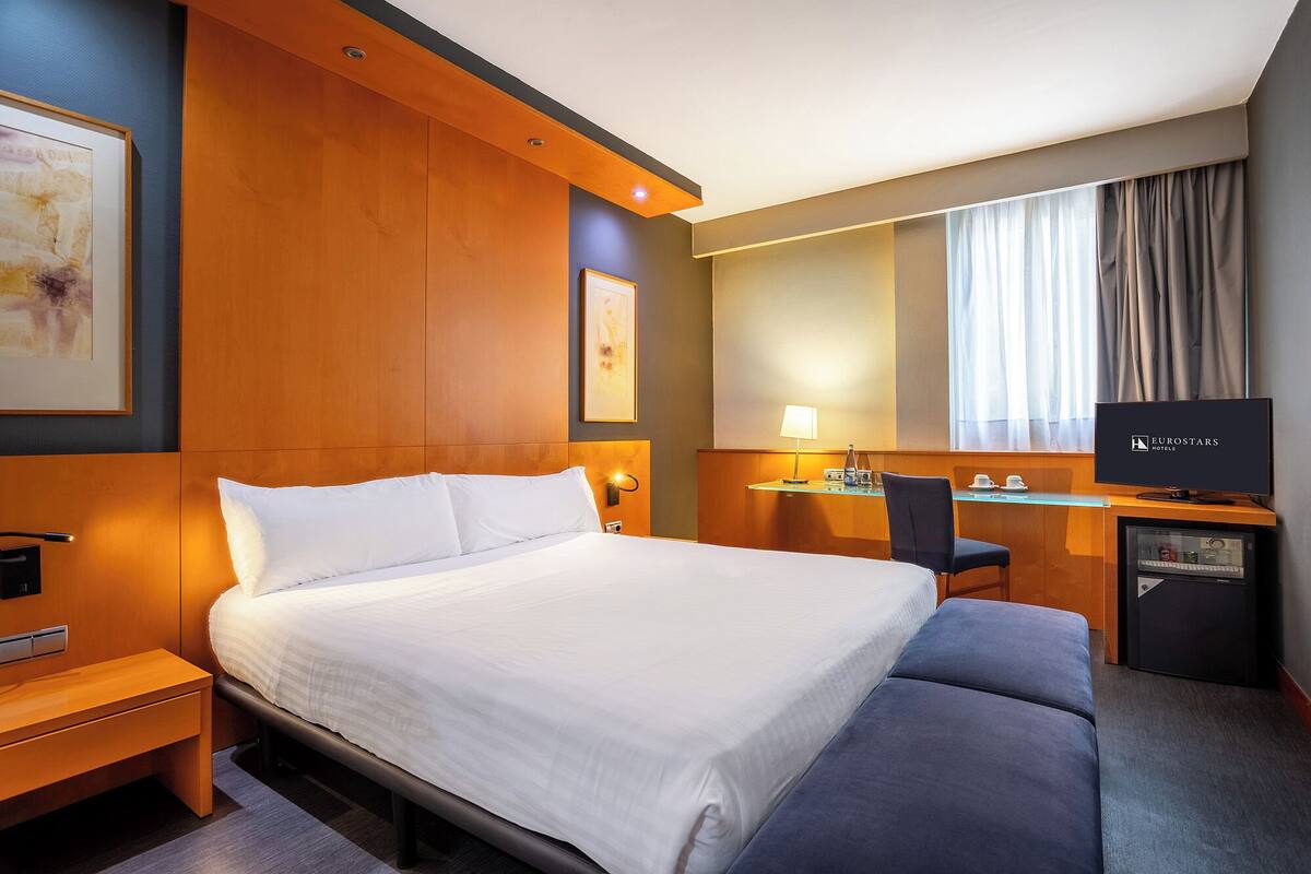 Een hotelkamer met een groot bed, een bureau met een computer, een televisie en een raam met gordijnen.