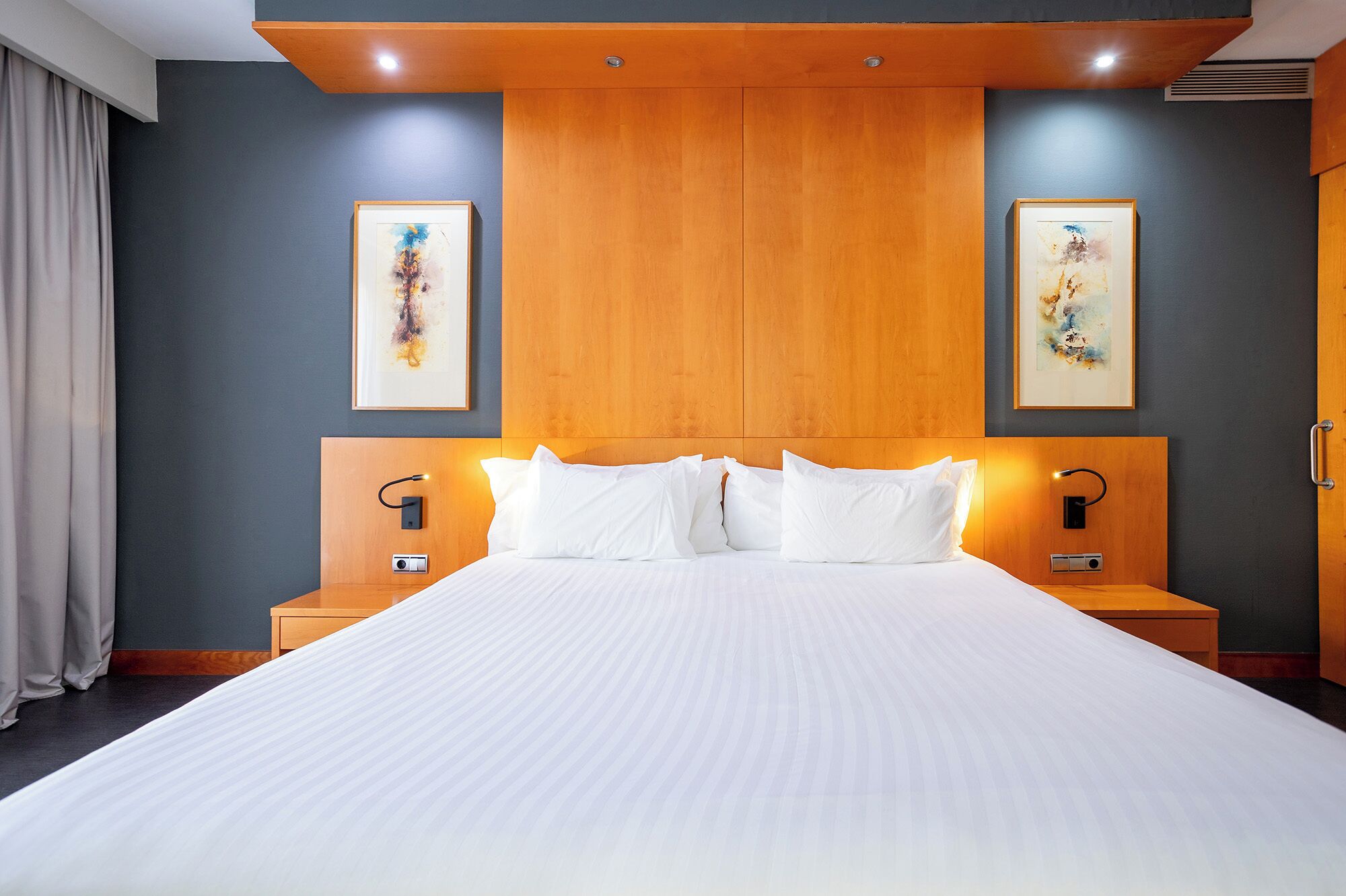 A hotel room with a large bed, wooden headboard, two bedside tables with lamps, and two framed artworks on the walls.