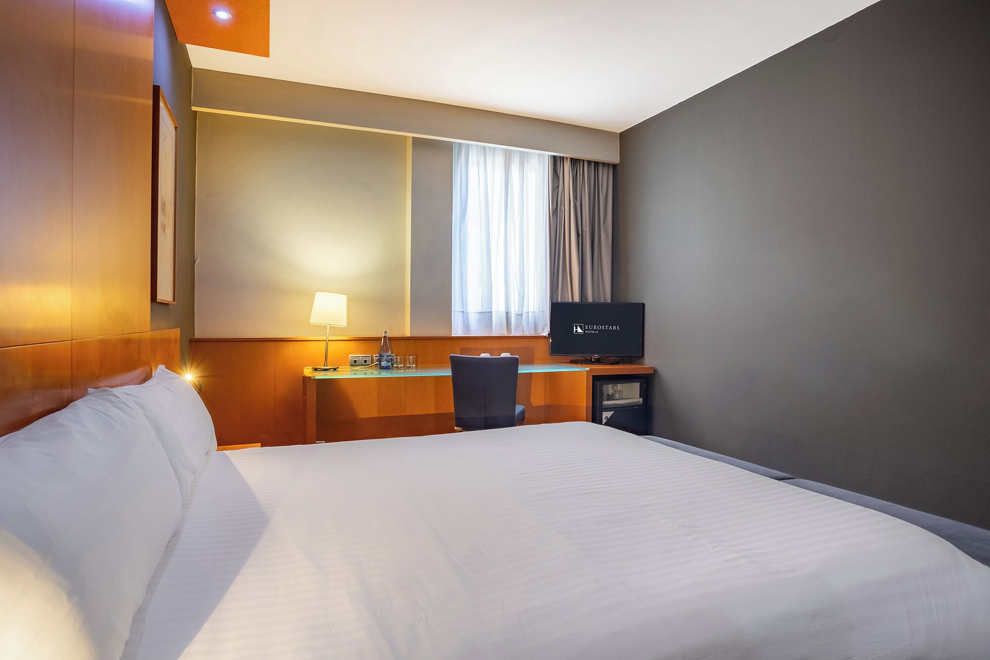 Una habitación de hotel con una cama, un escritorio con lámpara, un televisor y una ventana con cortinas.