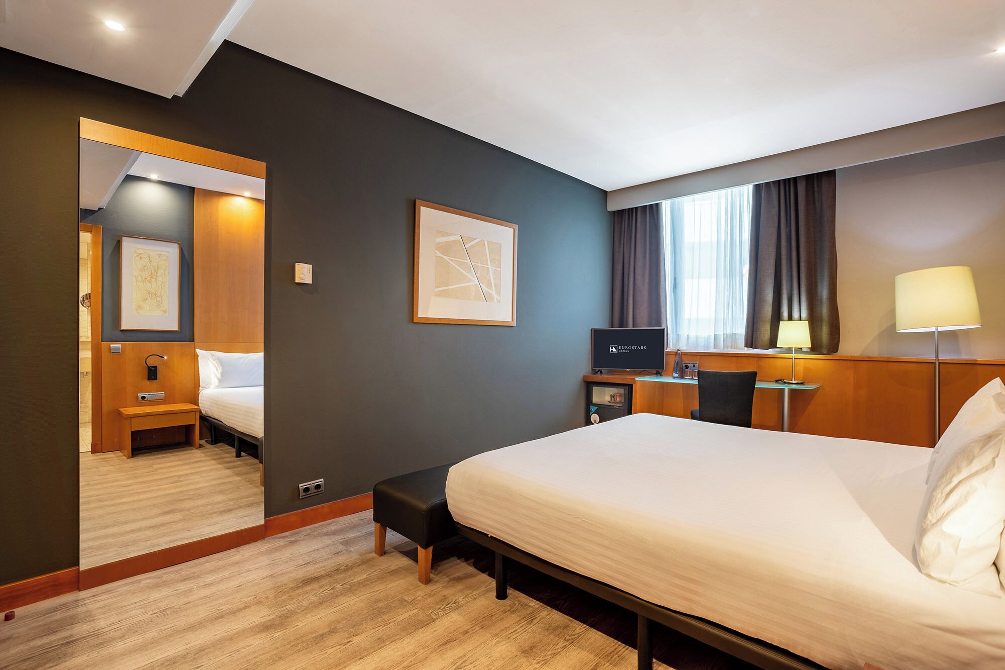 A hotel room with a large bed, a desk with a computer, a TV, and a mirror.