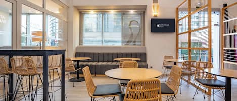 A modern lounge area with round tables and wicker chairs, a sofa, and a TV screen displaying the hotel name.