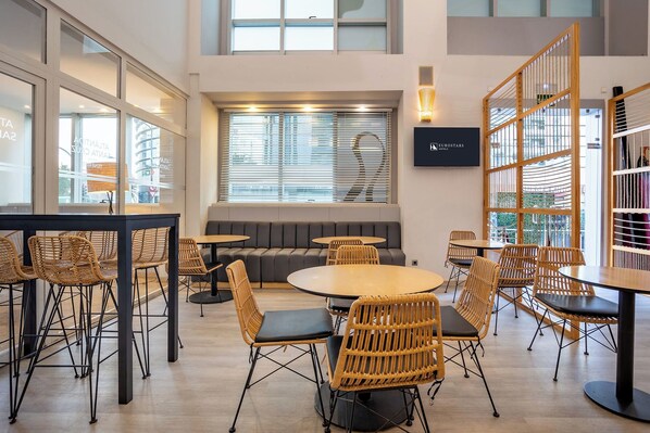 A modern lounge area with round tables and wicker chairs, a sofa, and a TV screen displaying the hotel name.