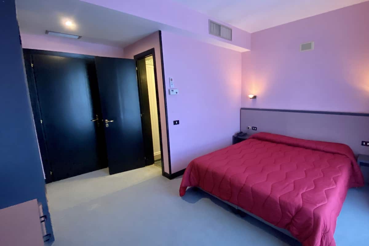 A hotel room with a bed, a door, and a wall-mounted light fixture.
