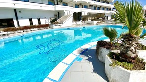 A swimming pool with a clear blue water, surrounded by a white building with multiple balconies and air conditioning units. - Hotel Clorinda (Capaccio-Paestum)