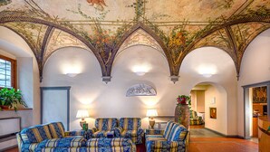 Una habitación con un techo alto y ornamentado que presenta un diseño floral y geométrico, una zona de estar con sofás estampados, y vistas a un pasillo con una puerta y una planta.