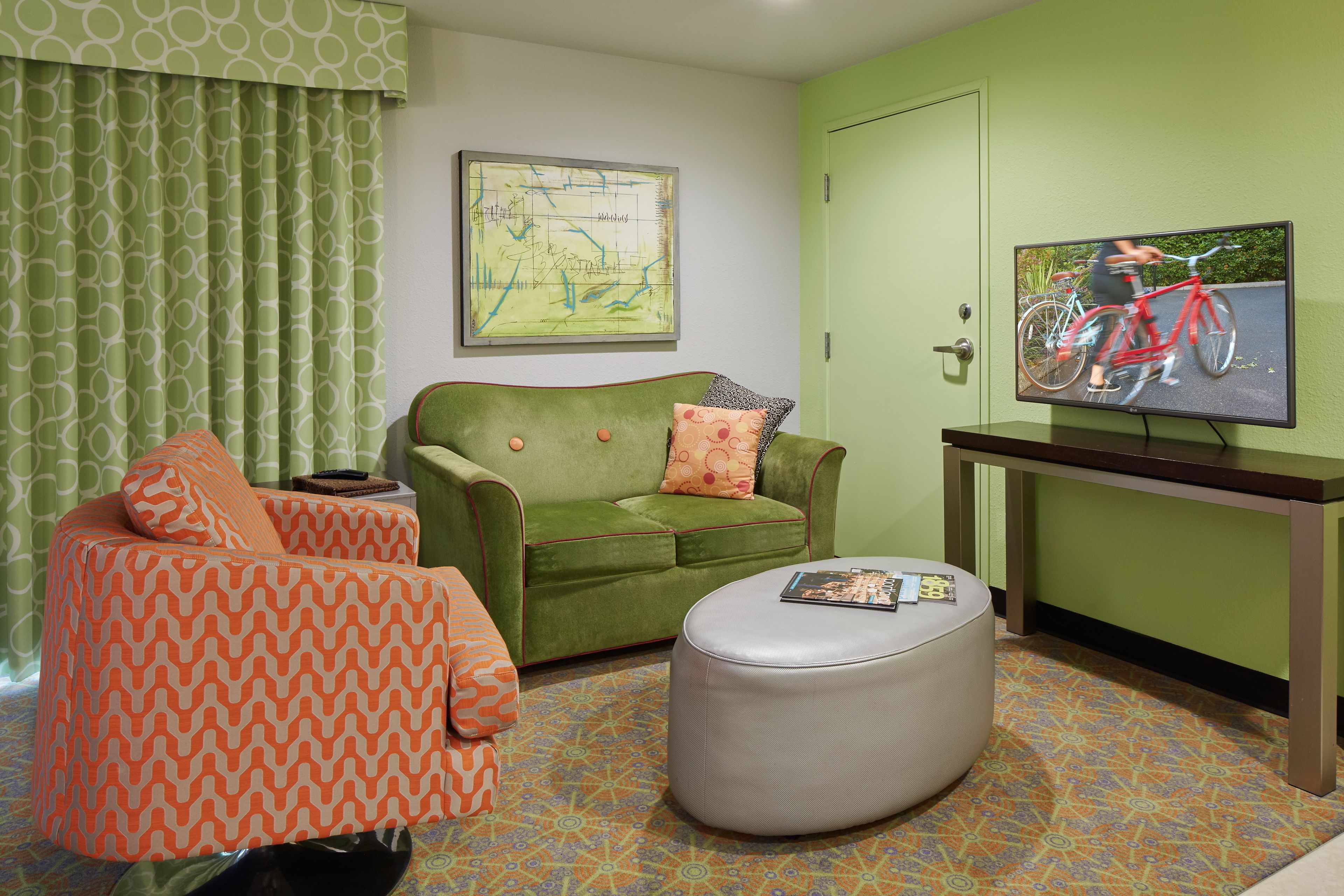 Un salon avec un canapé vert, un fauteuil à motifs orange, un pouf gris, une télévision et un sol à motifs.