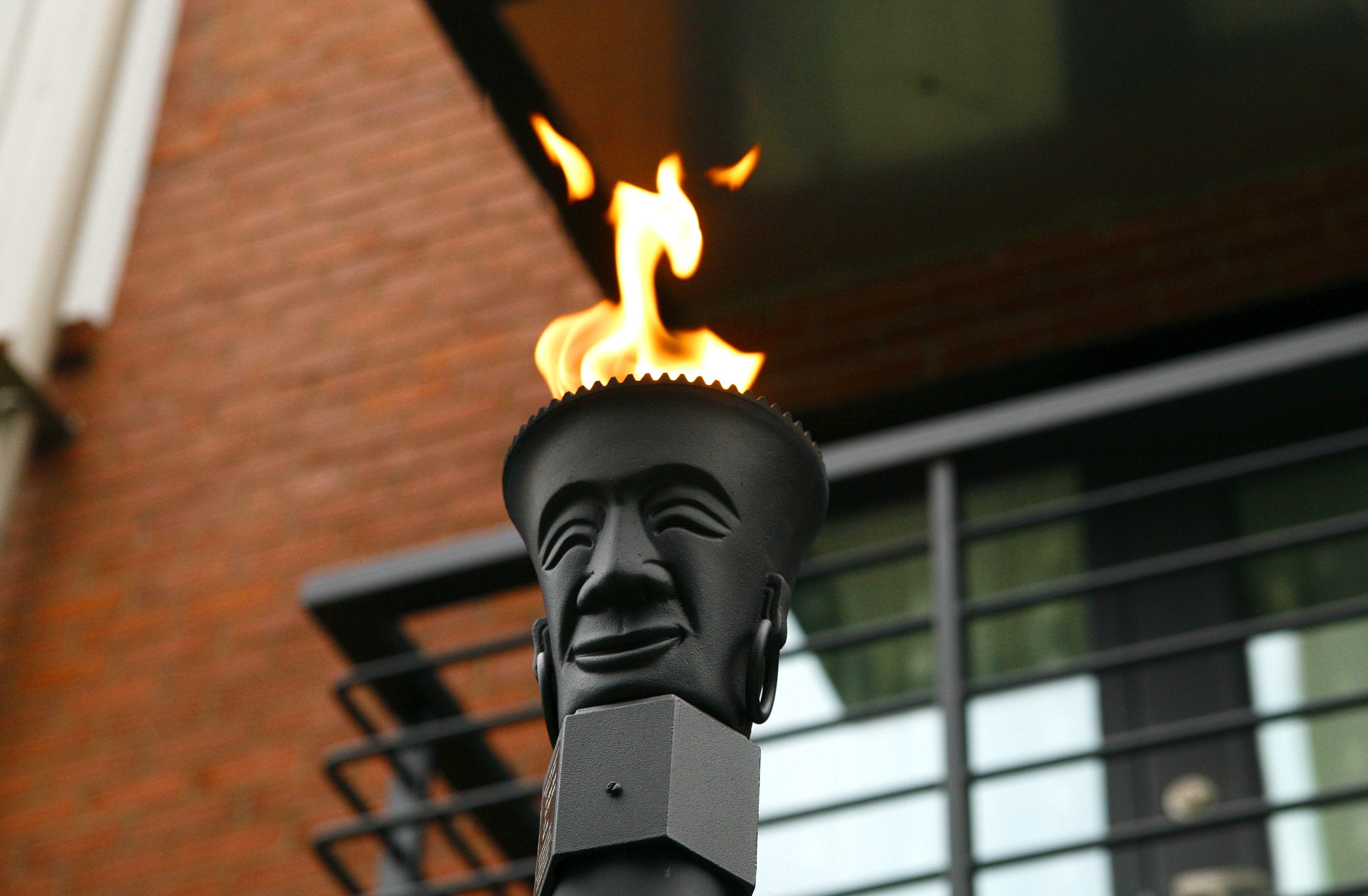 Uma estátua com uma chama no topo, diante de um muro de tijolos e uma janela com grades de metal.