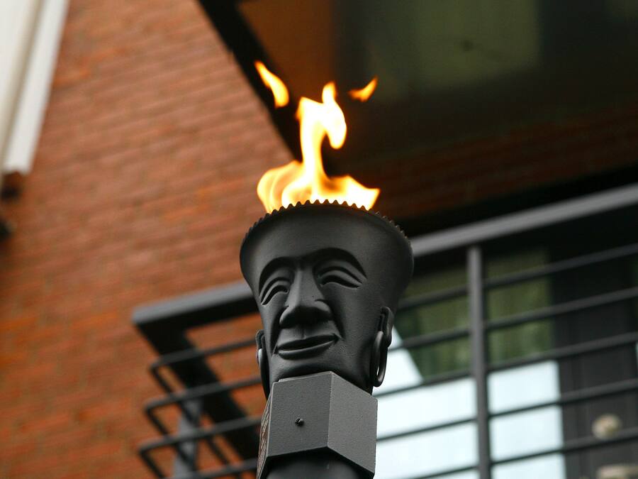Uma estátua com uma chama no topo, diante de um muro de tijolos e uma janela com grades de metal.