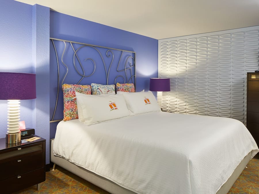 A neatly made bed with white bedding, a metal headboard, and colorful pillows. There are two bedside tables with lamps, a dresser, and a wall-mounted TV.