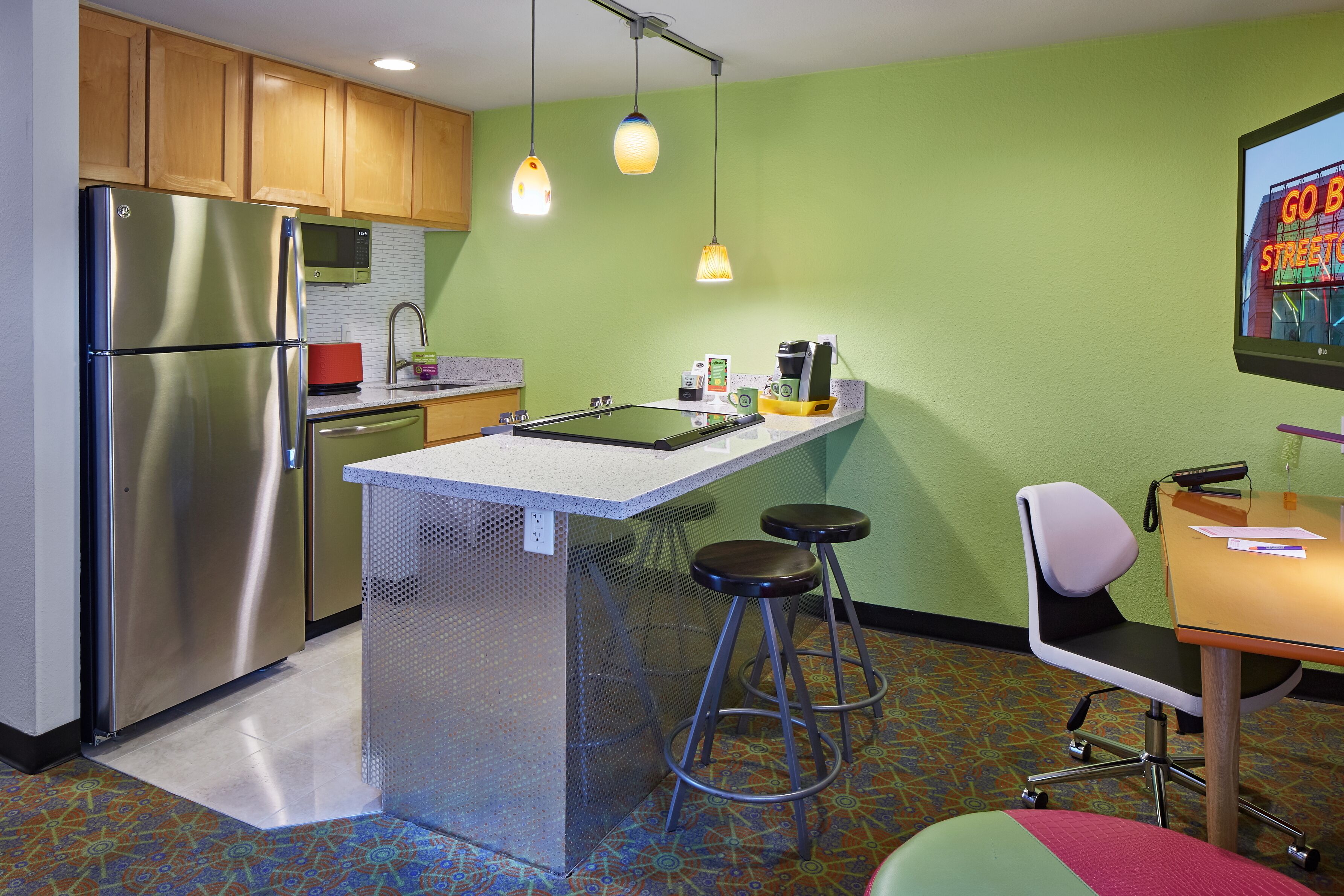 A compact kitchen with a stainless steel refrigerator, a microwave, and a sink. There are two stools and a small table with a chair. The walls are painted green, and there is a patterned floor.