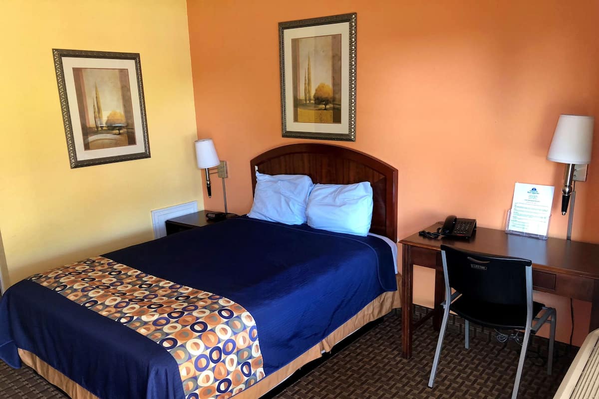 Una habitación de hotel con una cama, un escritorio, dos sillas, una lámpara y dos cuadros en la pared.