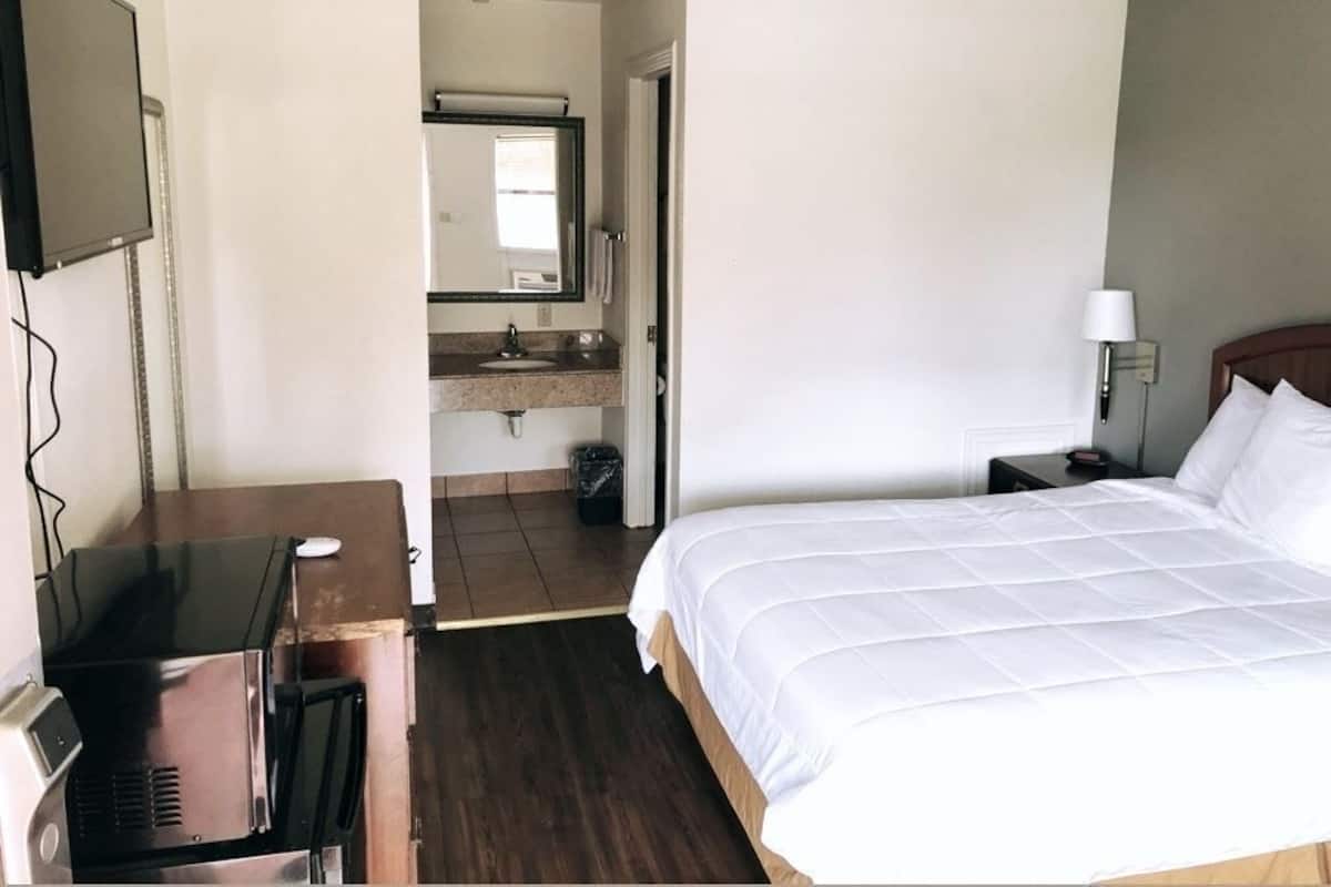 Habitación de hotel con cama, escritorio con computadora, televisor, baño con lavamanos y espejo, y mesita de noche con lámpara.