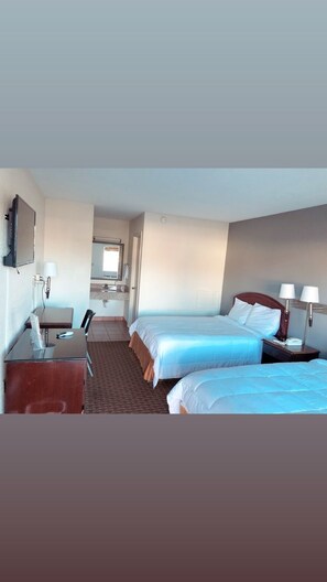 A hotel room with two beds, a desk with a chair, a small table, a mirror, and a bathroom visible through an open door.