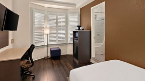 A hotel room with a bed, a desk with a chair, a television, a window with blinds, a shower, and a refrigerator. - Mithila San Francisco, SureStay Collection by Best Western (San Francisco)