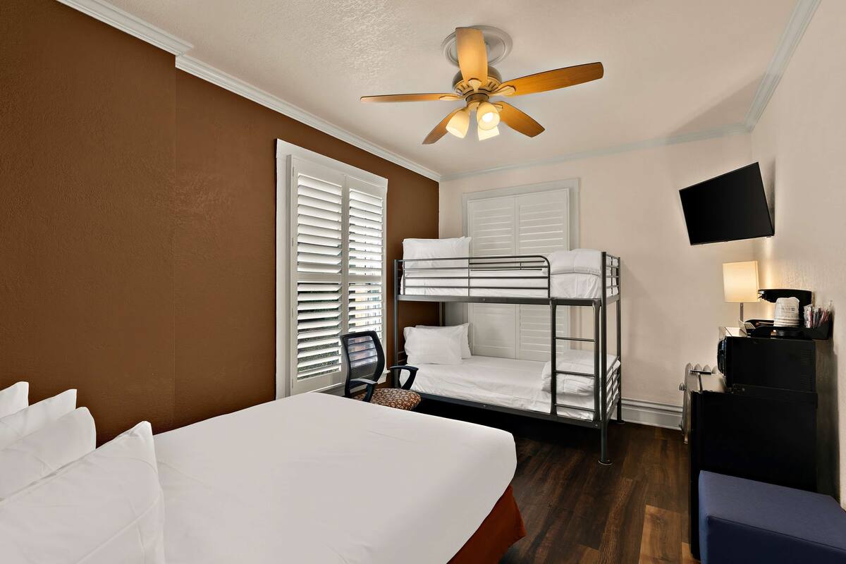 A hotel room with a bunk bed, a single bed, a ceiling fan, a TV mounted on the wall, and a desk with a lamp.