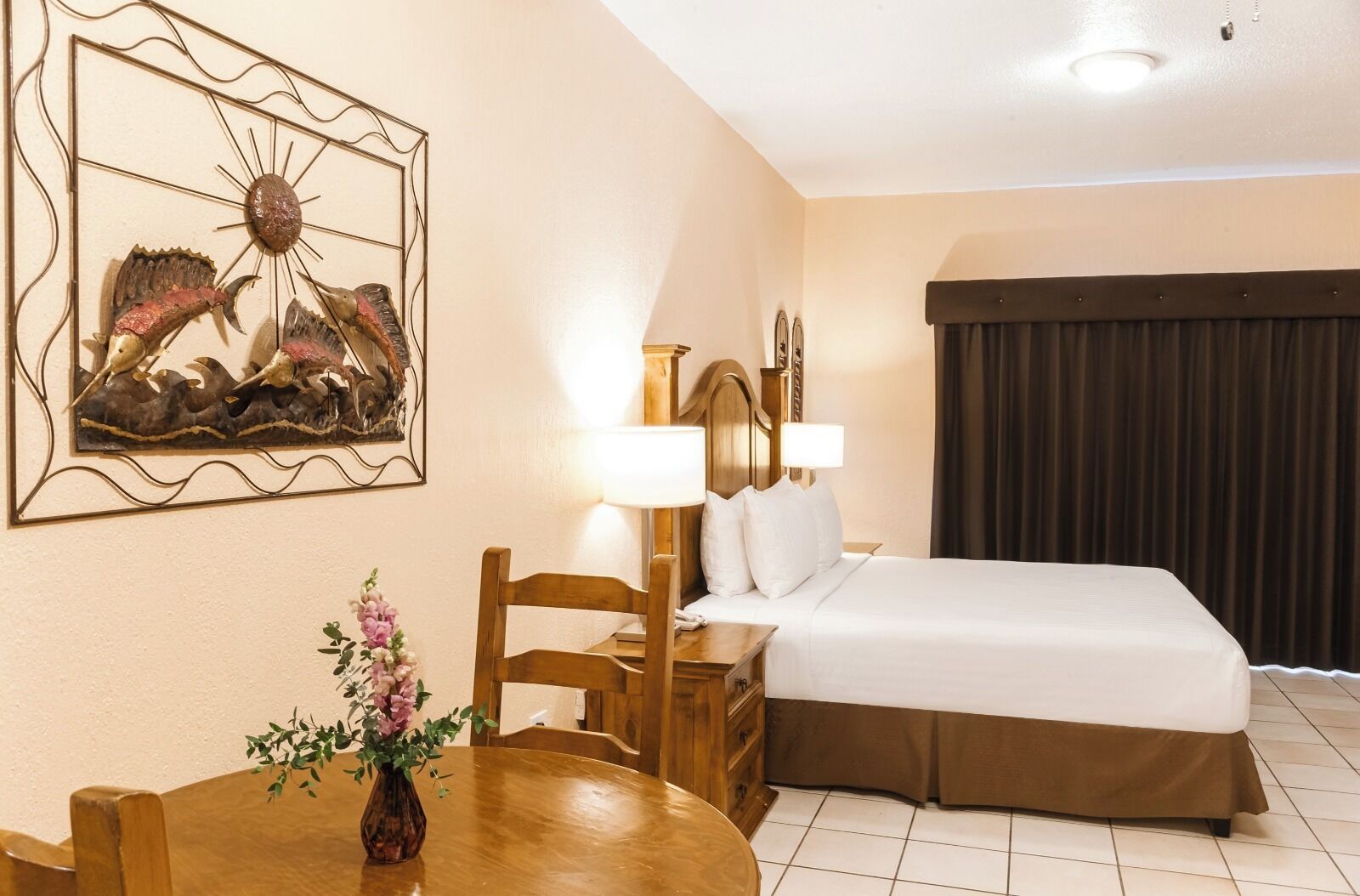 A hotel room with a bed, a desk with a lamp, a chair, a painting on the wall, and a window with curtains.