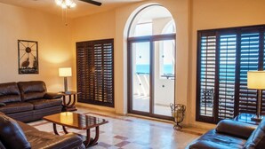 Sala de estar con sofá, sillón y mesa de centro, un ventilador de techo y vistas al océano a través de amplios ventanales.