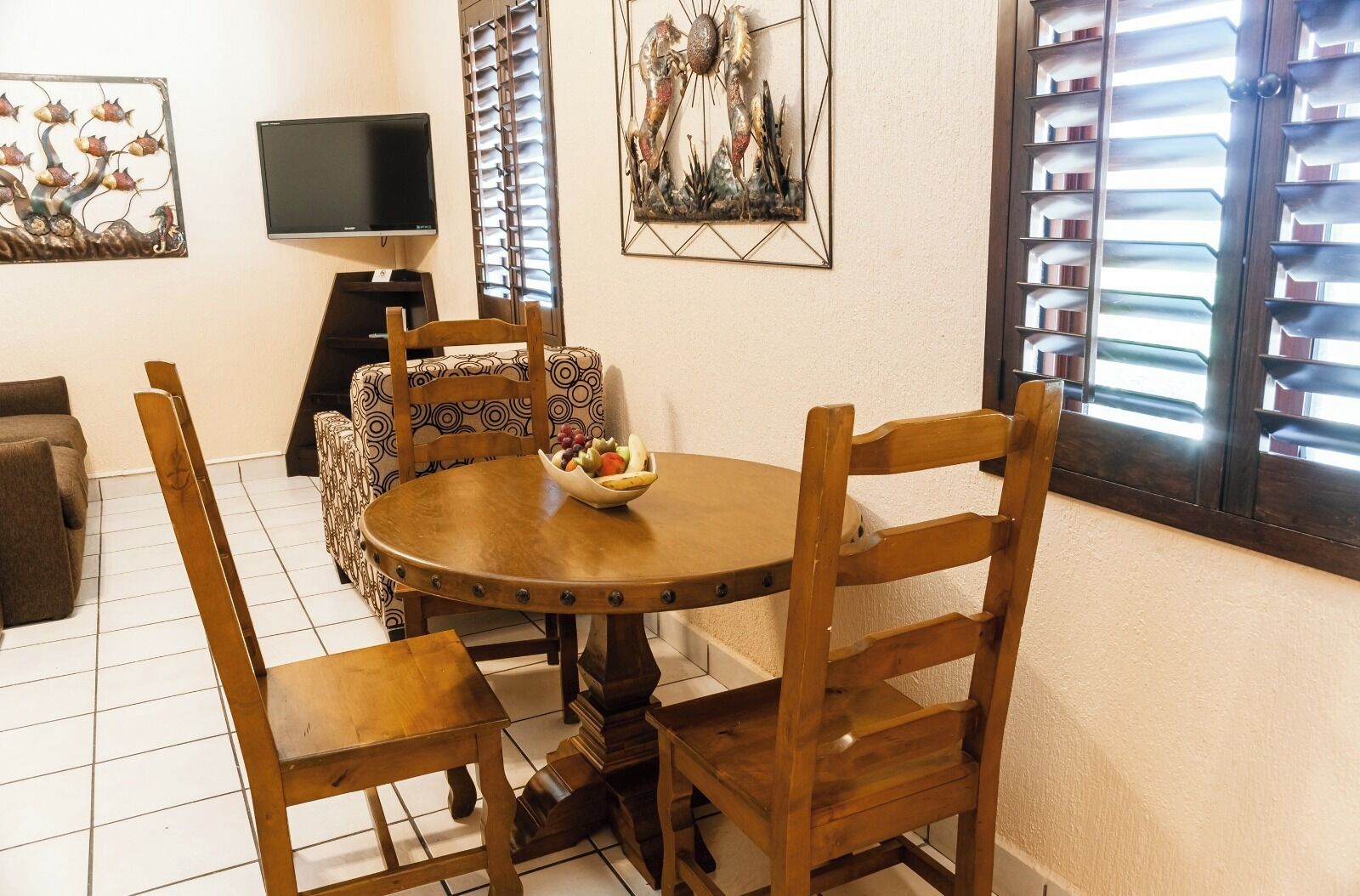 Un comedor con una mesa redonda de madera, cuatro sillas, un frutero, un televisor y obras de arte en las paredes.