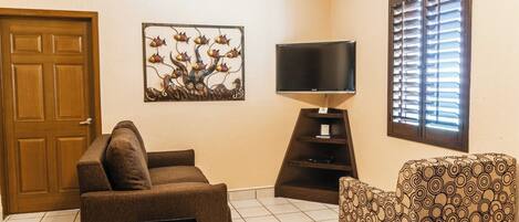 A living room with a brown sofa, a patterned armchair, a flat-screen TV, a decorative wall piece, and a window with blinds.