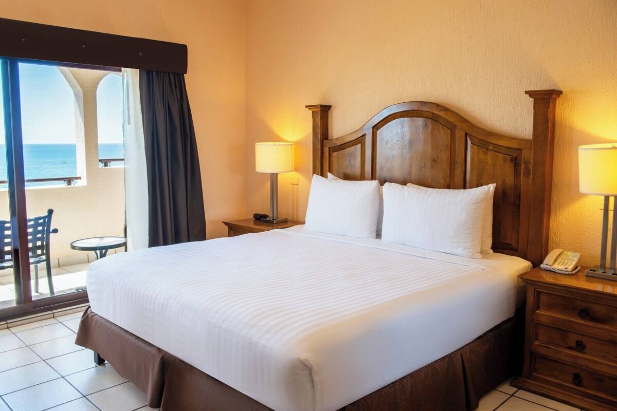 Habitación de hotel con una cama grande, dos mesitas de noche con lámparas, un cabecero de madera y vistas al mar desde un balcón.