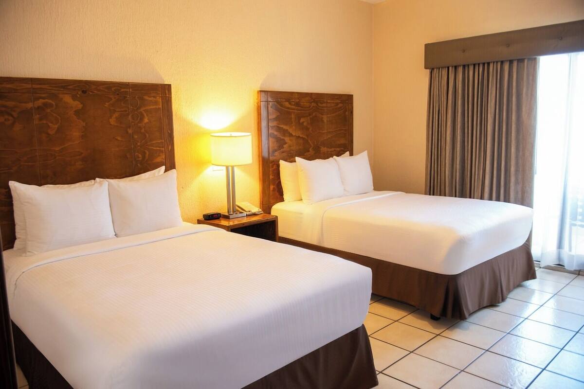 Habitación de hotel con dos camas, una mesita de noche con lámpara y una ventana con cortinas.