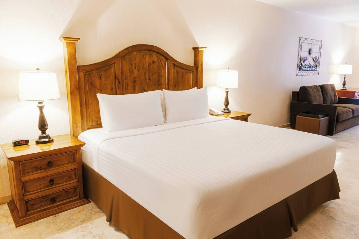 Una cama bien hecha con tablero de madera, dos mesitas de noche con lámparas, un pequeño atril y un sofá en una habitación de hotel.