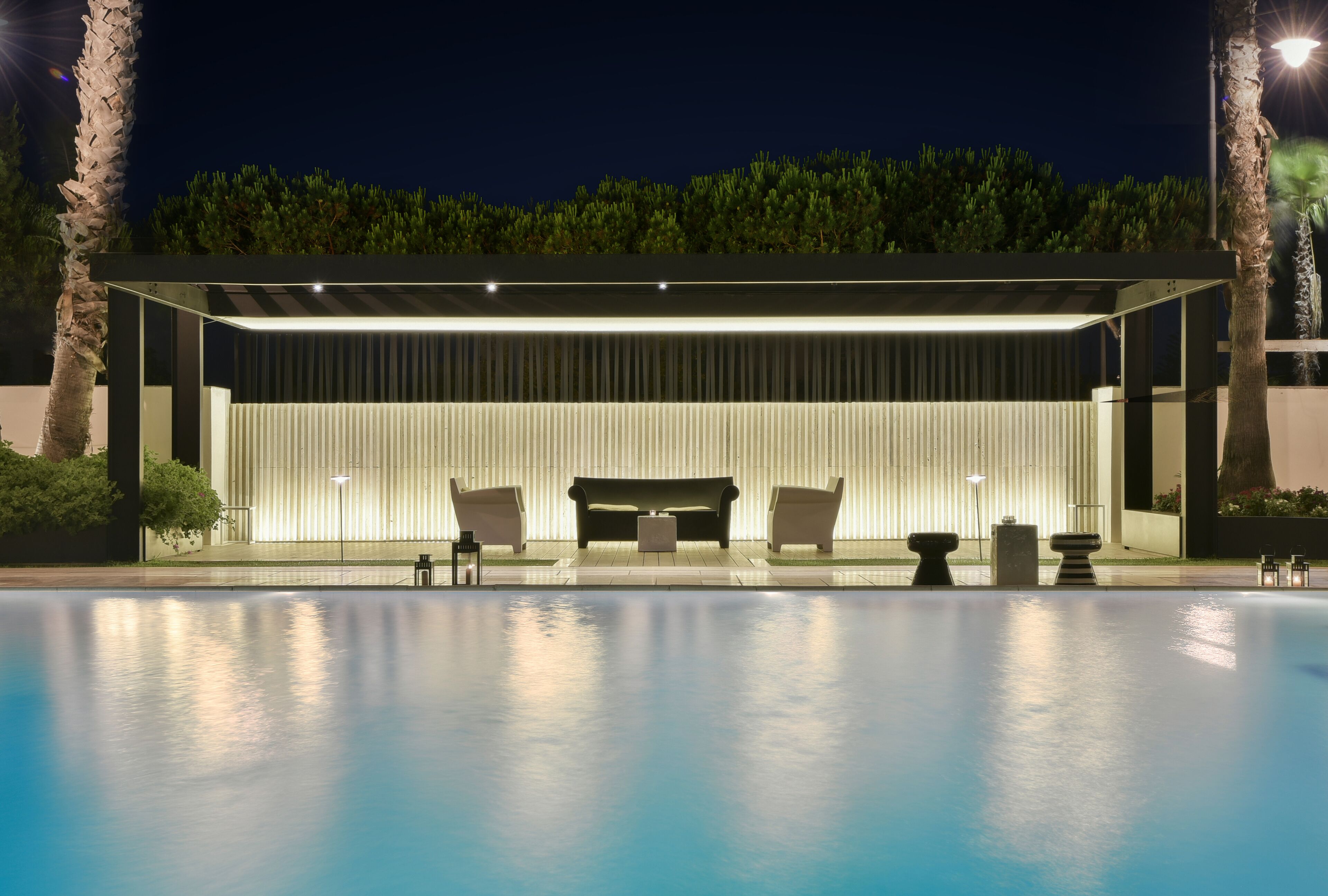 A modern outdoor pool area with a covered seating area, a pool, and surrounding greenery.