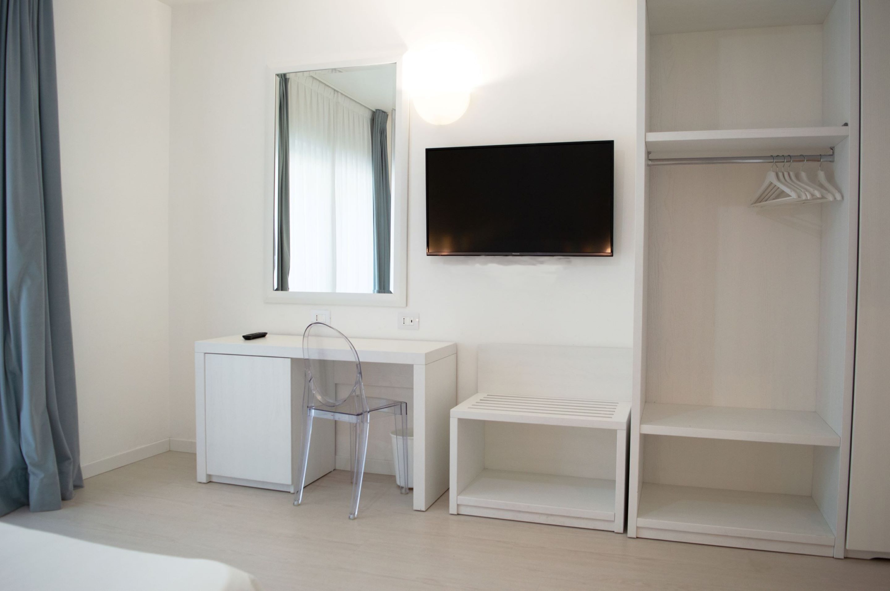 Ein modernes, minimalistisches Schlafzimmer mit weißem Schreibtisch, einem Sessel, einem Fernseher und einem begehbaren Kleiderschrank.