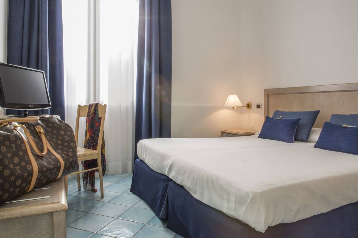 A hotel room with a bed, a desk with a computer monitor, a chair, a lamp, and a suitcase.