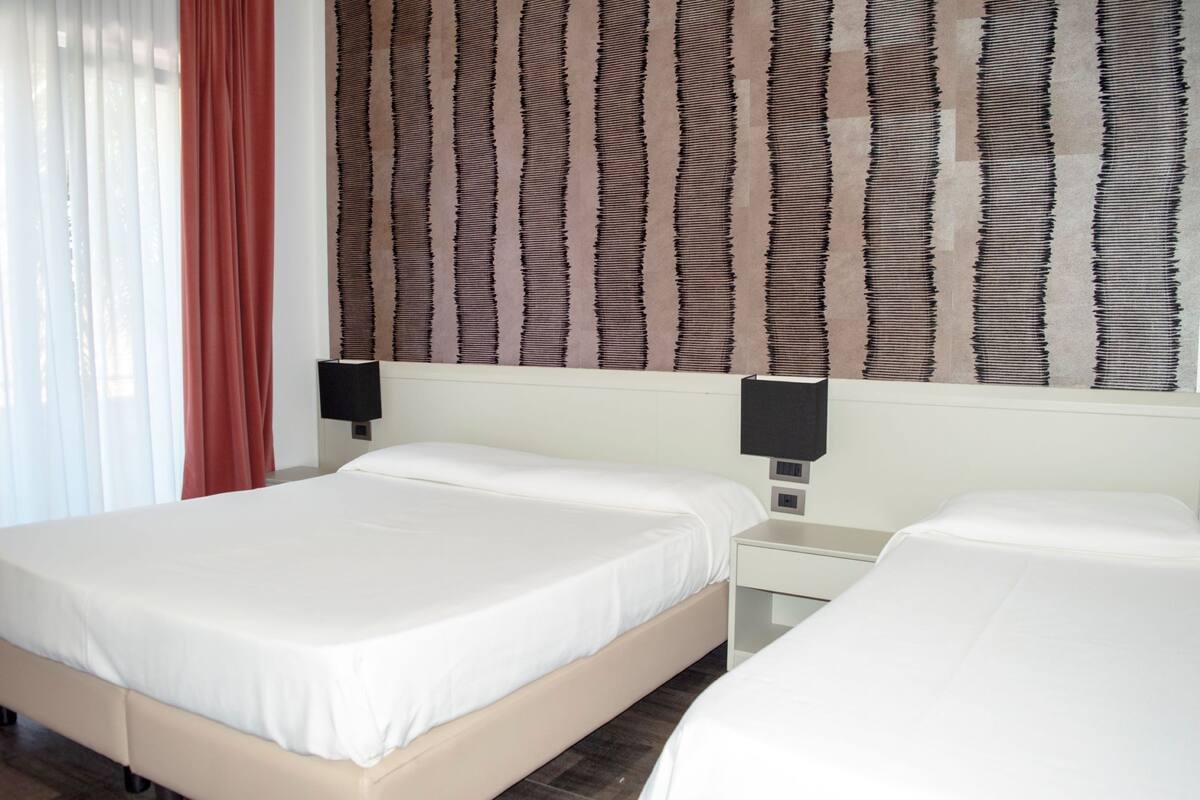 A hotel room with two beds, a large window with sheer curtains, a wall with a textured pattern, and two bedside tables with lamps.