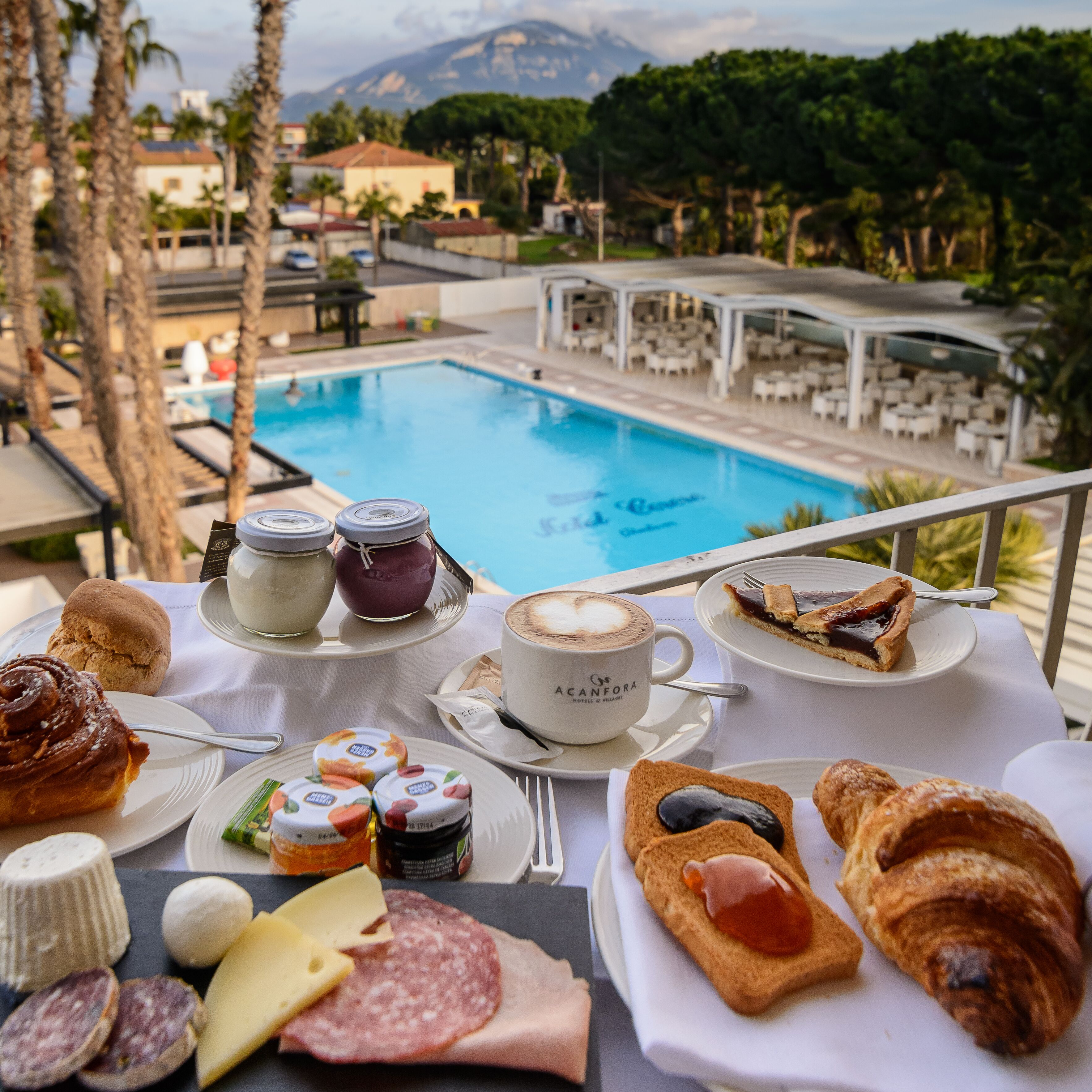 Una mesa con alimentos para el desayuno, como pasteles, queso y una taza de café, con vista a una piscina y un área de descanso.