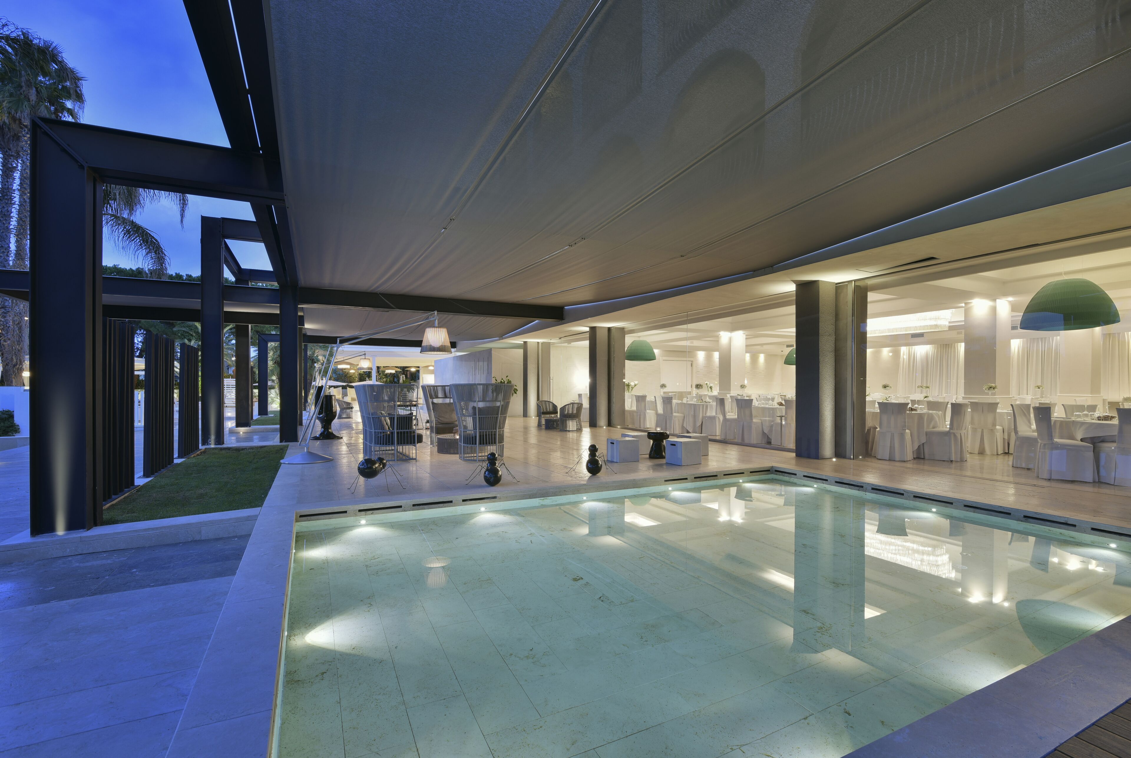 A modern indoor pool area with clear glass walls, a pool, and seating areas with tables and chairs.