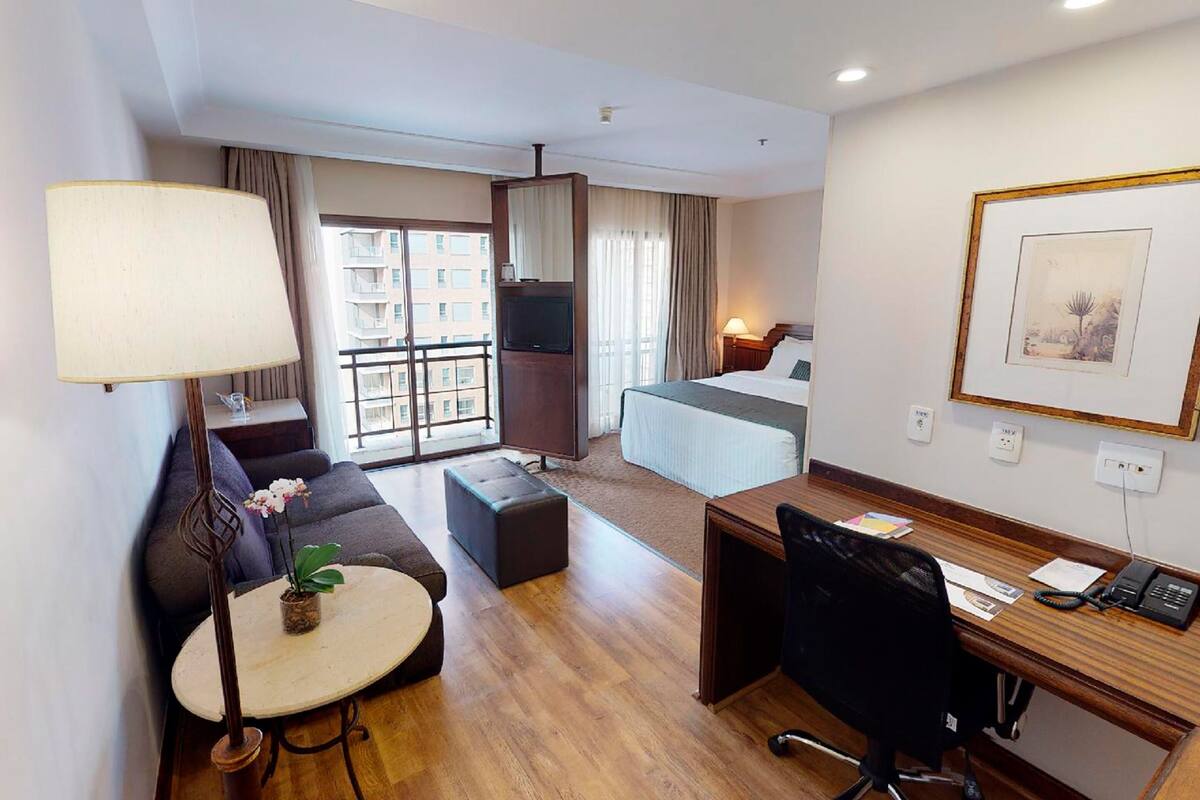 A hotel room with a sofa, a desk with a chair, a small table with a lamp, a bed, and a TV.