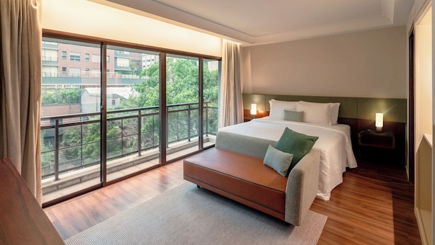 A modern hotel room with a large window, a bed with white linens, a wooden bench, and a nightstand with a lamp. - Estanplaza Paulista (São Paulo)