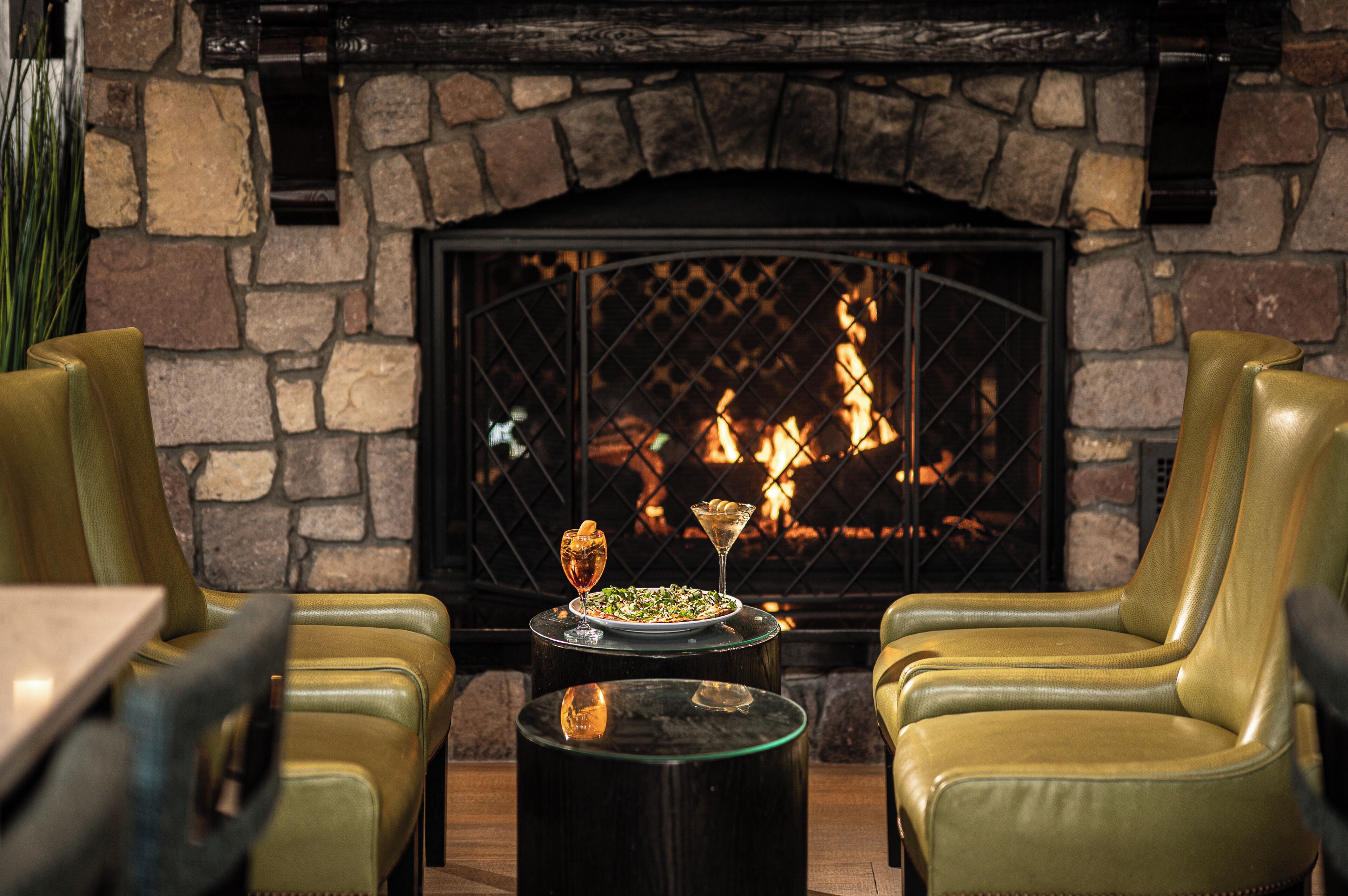 A cozy seating area with leather chairs and a table, a fireplace with a stone wall backdrop, and a plate of food with drinks on the table.