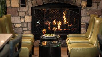 A cozy seating area with leather chairs and a table, a fireplace with a stone wall backdrop, and a plate of food with drinks on the table.
